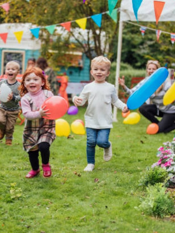 Three young children enjoy playing with balloons at a garden party as they run towards the camera. Adults are sat talking in the background and looking at the children. There is a food truck in the background and a marquee and colorful bunting fill the garden.