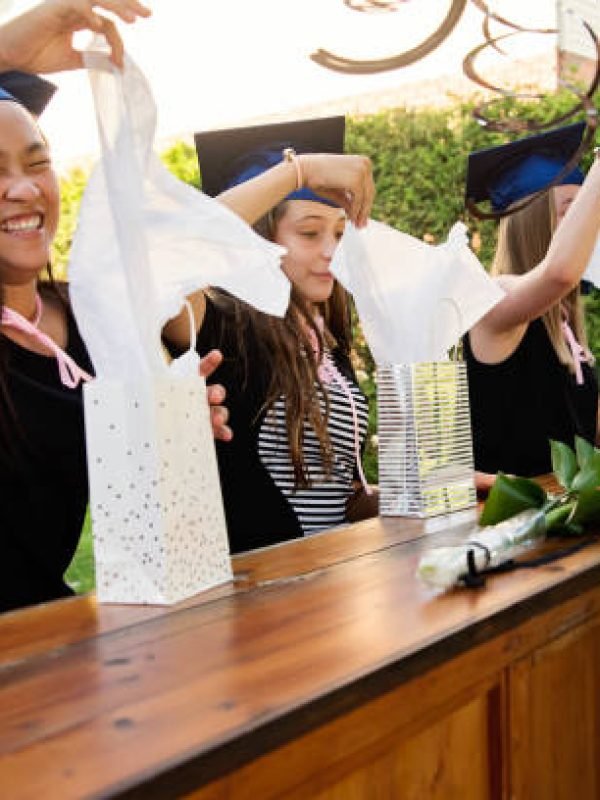 Celebrating primary school graduation in the backyard during Covid-19 pandemic for three multi-ethnic teenage girls.They are opening gifts wearing their graduation hats sitting on terrasse. Horizontal outdoors waist up shot with copy space.