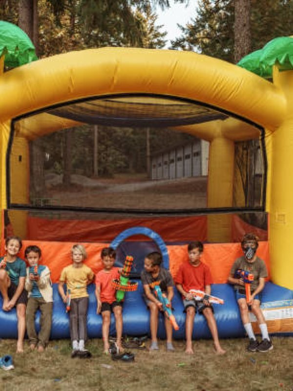 A multiracial group of elementary age and tween boys sit in a row along the edge of an inflatable bouncy castle and hold toy nerf guns while at a back yard birthday party.